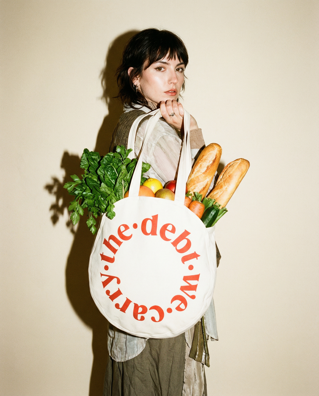 Person holding a tote bag with groceries and 'the debt we carry' text against a beige background
